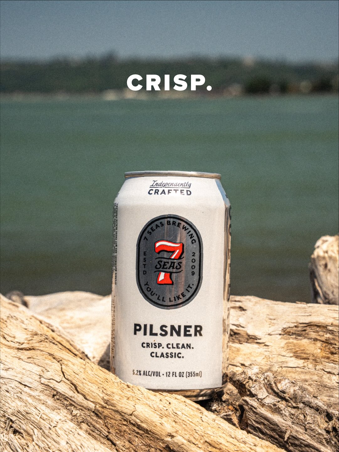 A can of 7 Seas Pilsner on driftwood at the waterfront with the word CRISP above