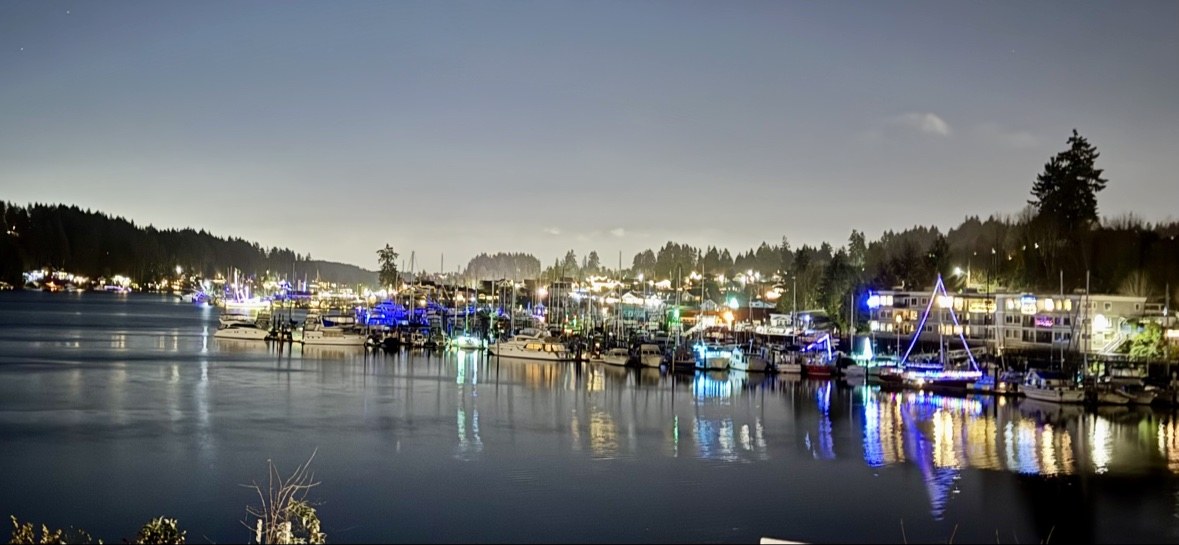 Boats decorated with holiday lights on the Gig Harbor waterfront at night, reflected in the water